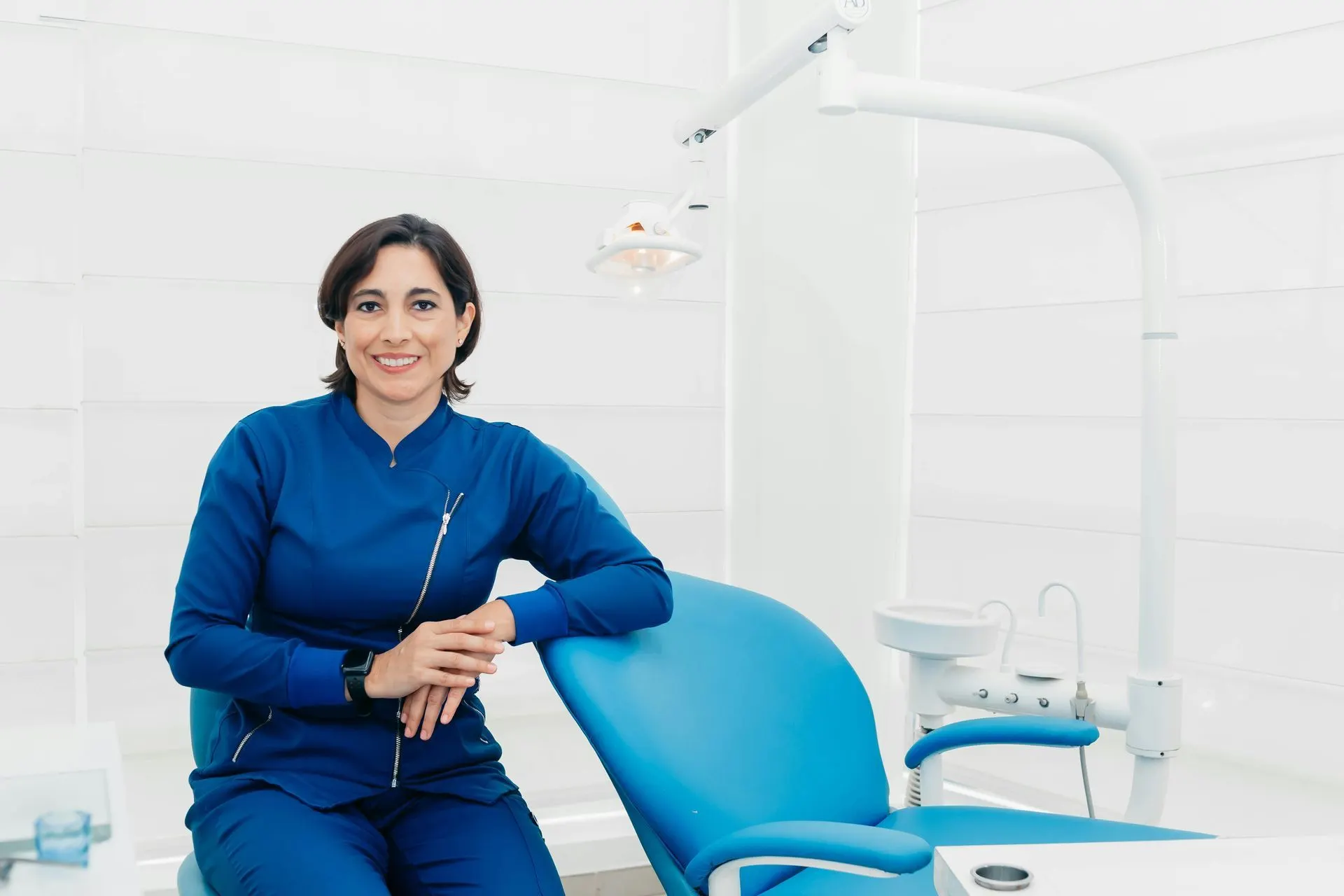 Dentist in blue scrubs seated beside a dental chair